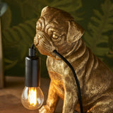 Endon Charthurst table lamp with a gold resin pug figure holding a black cable and exposed filament E14 bulb, set against leafy green decor.