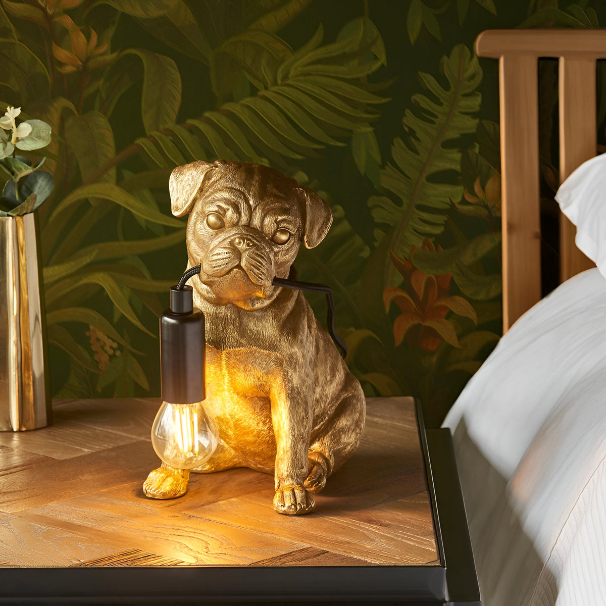 Endon Charthurst gold resin pug table lamp with black E14 bulb holder and exposed filament bulb, on a wooden bedside table against tropical leaf wallpaper.
