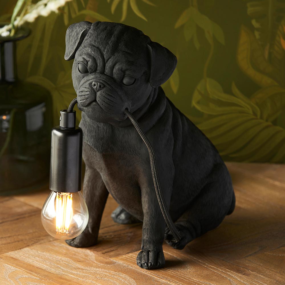 Endon Charthurst table lamp shaped as a matte black resin pug figure holding an E14 filament bulb on a fabric cable, set on wood against foliage wallpaper.