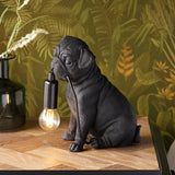 Endon Charthurst matt black resin pug table lamp with exposed E14 filament bulb on a wooden surface against tropical leaf wallpaper.
