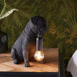 Endon Charthurst matt black resin pug table lamp with exposed E14 filament bulb, shown on a wooden bedside table against tropical leaf wallpaper.