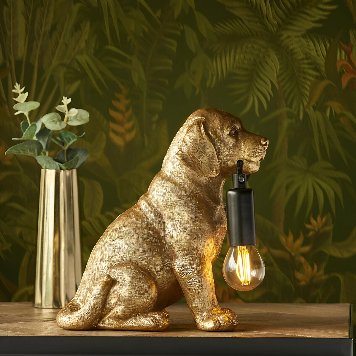 Brockworth Table Lamp in gold resin, shaped as a sitting Labrador puppy holding a black-fitted filament bulb, set against a leafy green backdrop.