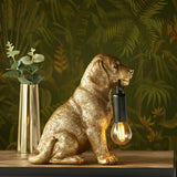 Brockworth Table Lamp in gold resin, shaped as a sitting Labrador puppy holding a black-fitted filament bulb, set against a leafy green backdrop.