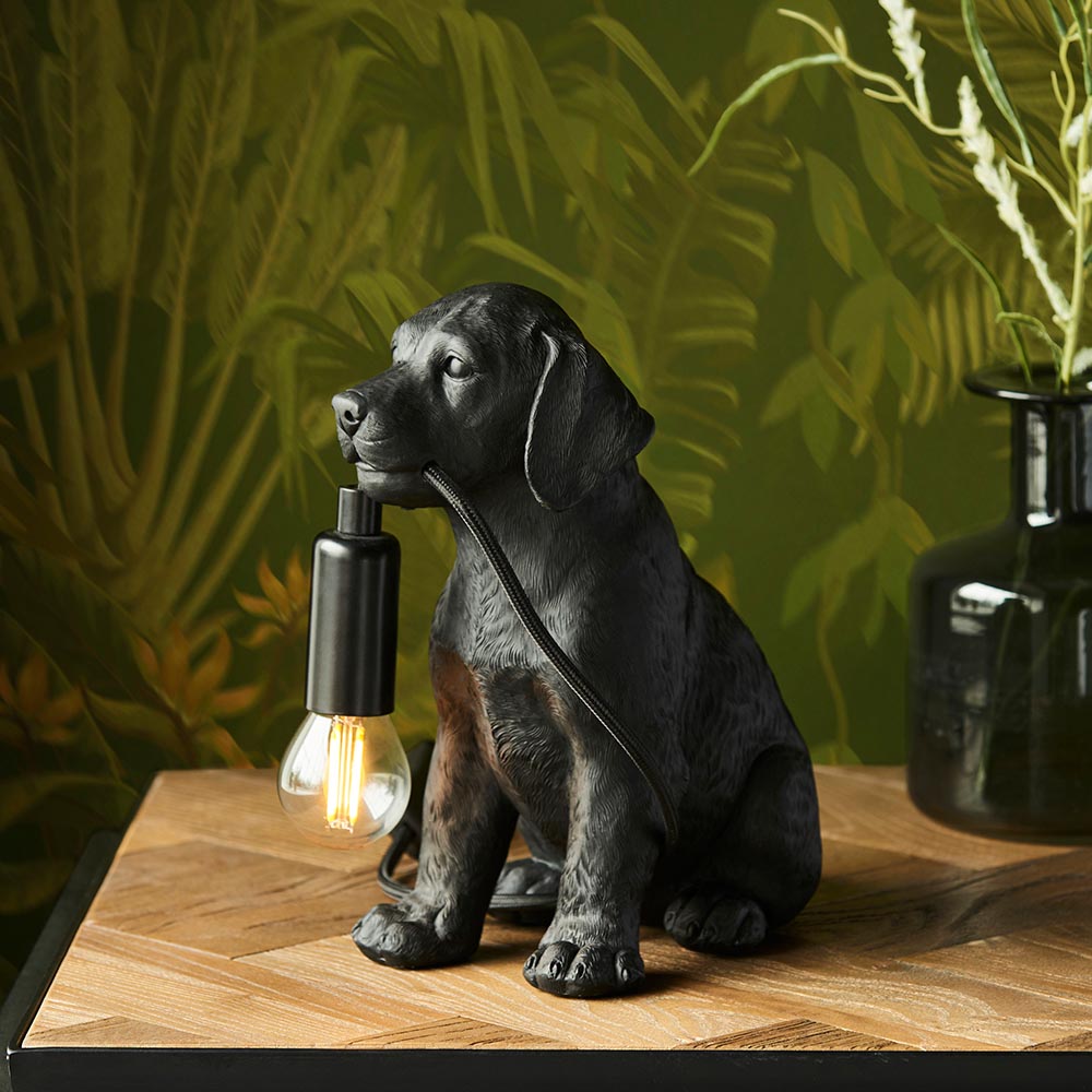 Endon Brockworth table lamp shaped as a matt black resin Labrador puppy holding an E14 filament bulb, on a wooden surface against leafy green wallpaper.