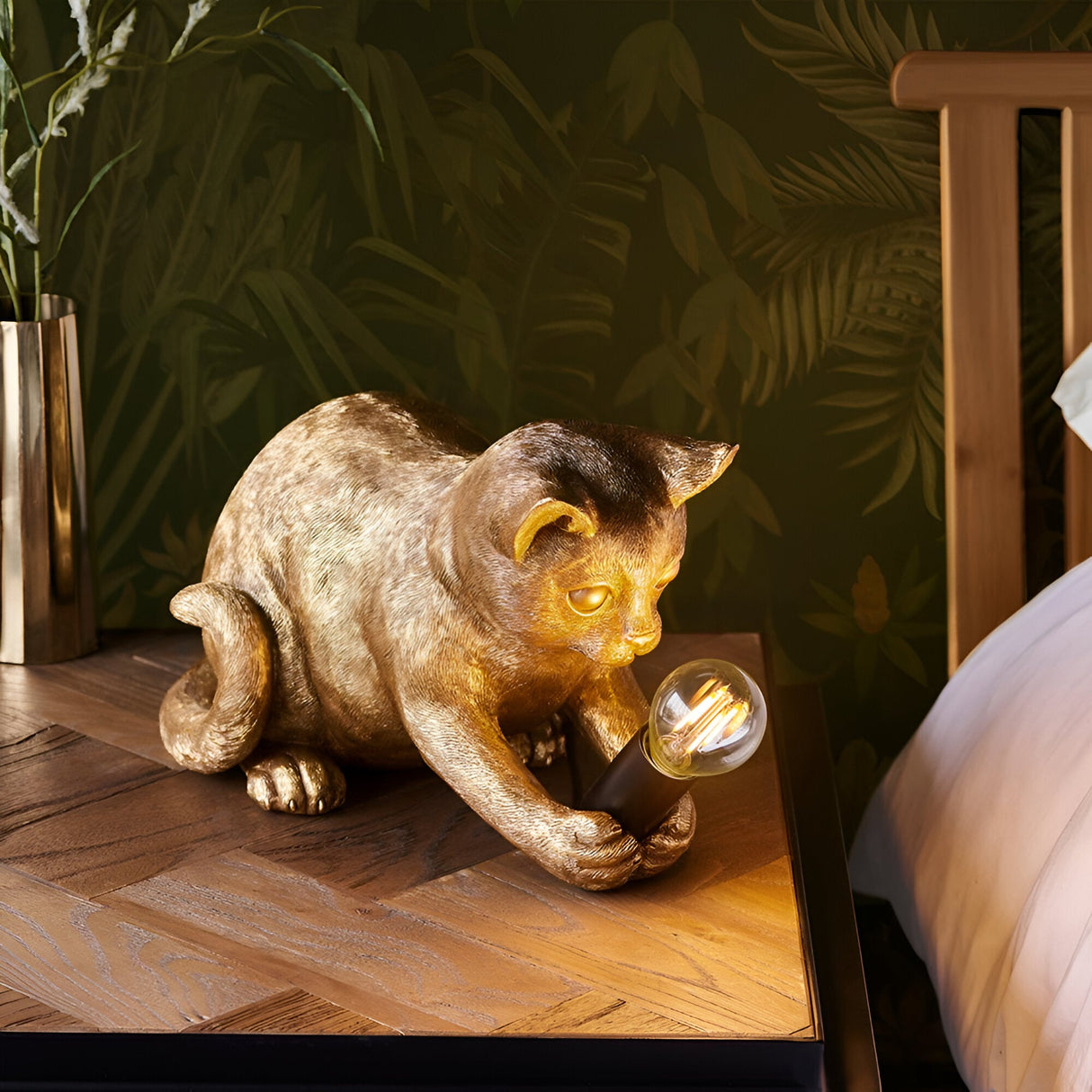 Endon Brockwick gold resin kitten table lamp with filament E14 bulb, shown on a wooden bedside table against tropical leaf wallpaper.