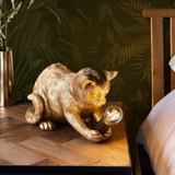 Endon Brockwick gold resin kitten table lamp with filament E14 bulb, shown on a wooden bedside table against tropical leaf wallpaper.