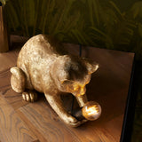 Endon Brockwick table lamp shaped as a crouching gold resin kitten holding a filament bulb in its paws, on a wooden surface with leafy backdrop.