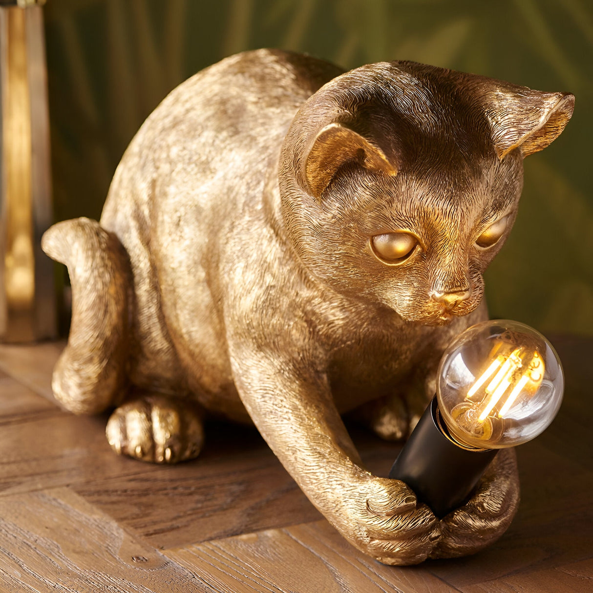 Endon Brockwick table lamp shaped as a gold resin kitten holding an exposed filament bulb in a matt black E14 holder, on a parquet floor.