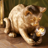 Endon Brockwick table lamp shaped as a gold resin kitten holding an exposed filament bulb in a matt black E14 holder, on a parquet floor.