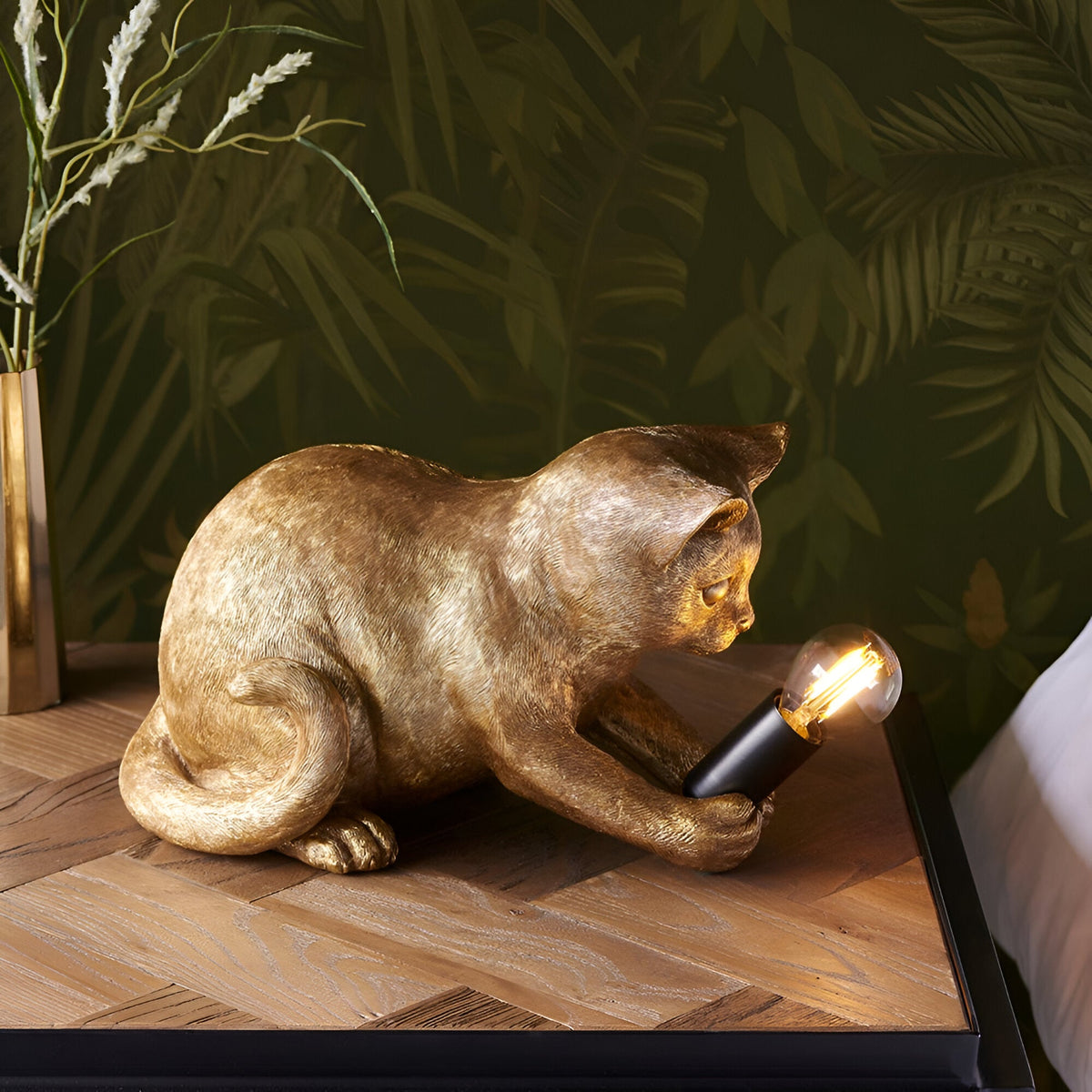Endon Brockwick table lamp shaped as a gold resin kitten holding a black E14 holder with filament bulb, on a wooden bedside table against palm wallpaper.