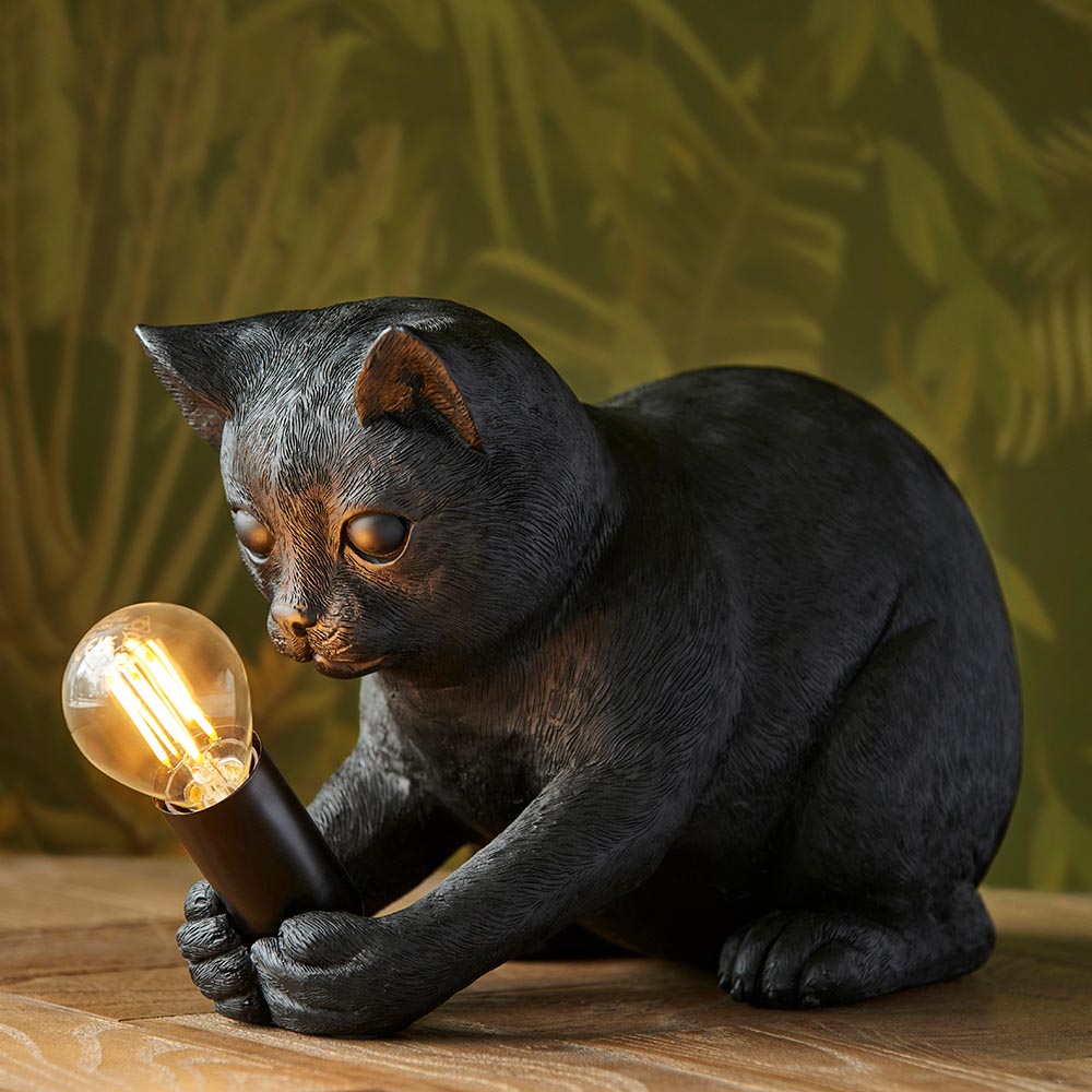 Endon Brockwick matt black resin kitten table lamp holding an E14 filament bulb, shown on a wooden surface against a leafy green backdrop.