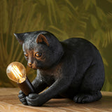 Endon Brockwick matt black resin kitten table lamp holding an E14 filament bulb, shown on a wooden surface against a leafy green backdrop.
