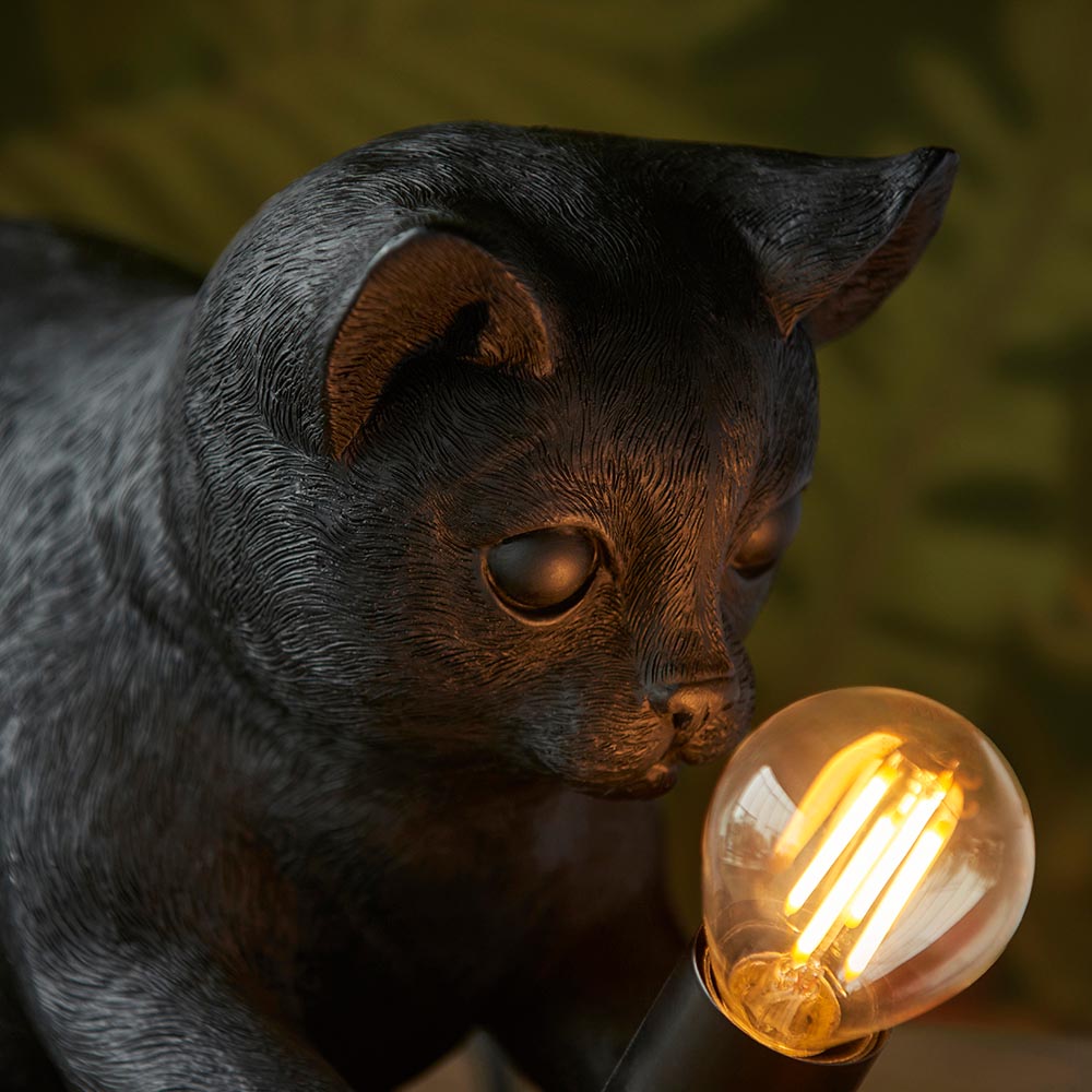 Endon Brockwick table lamp shaped as a matt black resin kitten, shown close-up sniffing a lit warm filament E14 bulb against a green leafy backdrop.