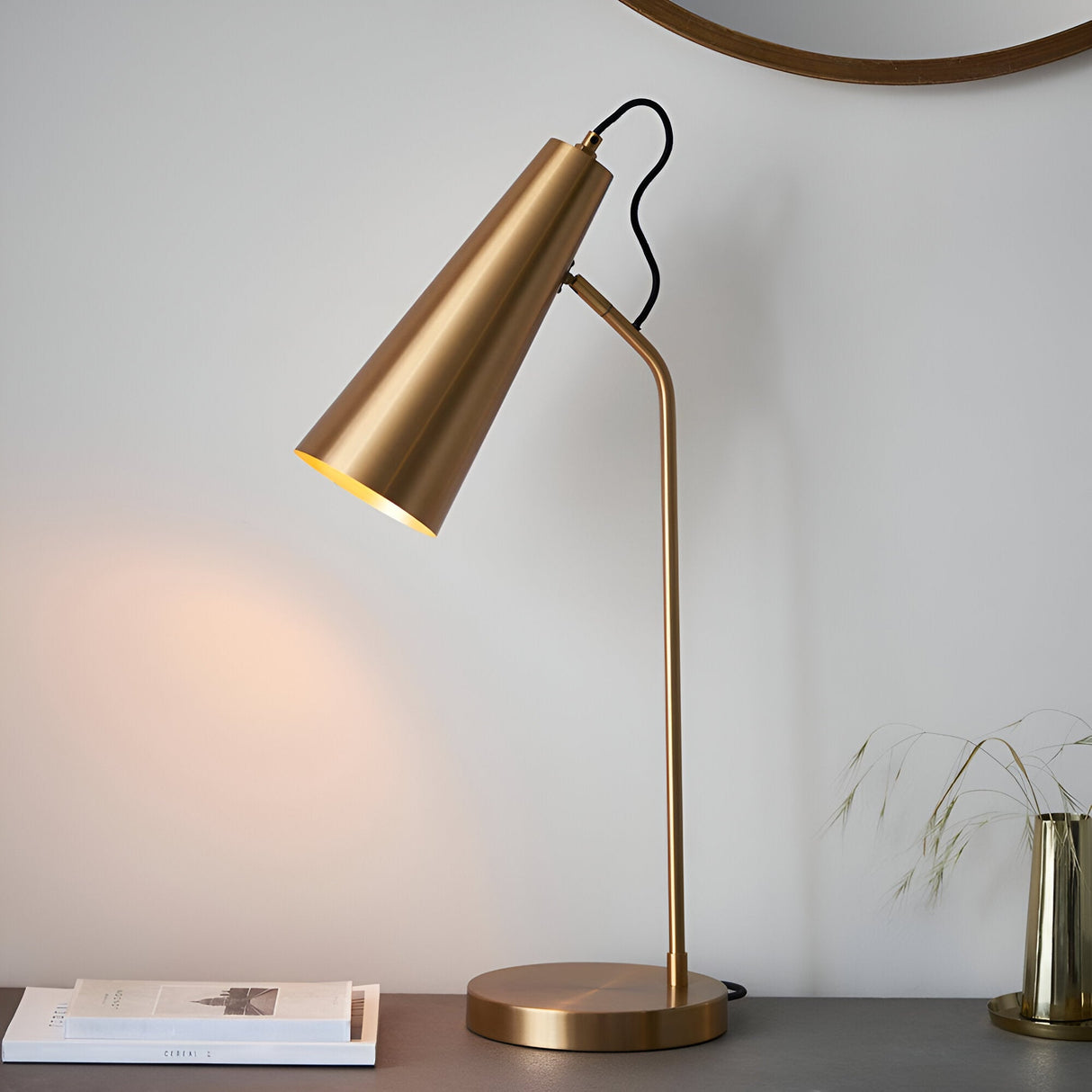 Endon Brockleigh antique brass table lamp with adjustable conical shade and black fabric flex, styled on a desk beside books and a round mirror.