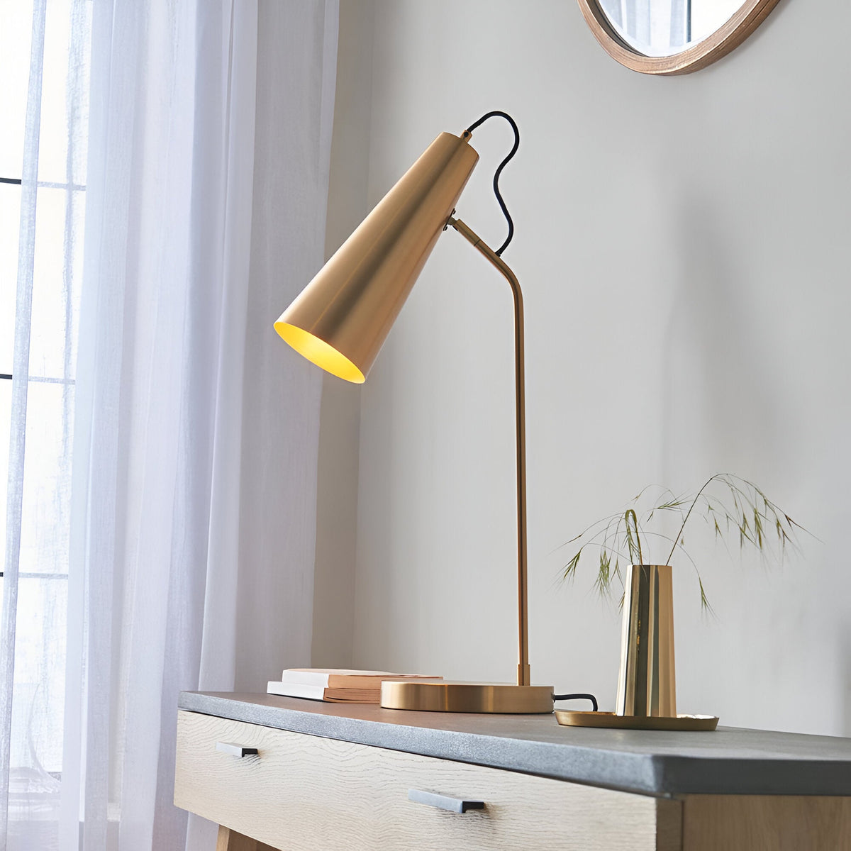 Endon Brockleigh antique brass table lamp with adjustable conical shade and black fabric flex, on a wooden console beside sheer curtains and a round mirror.