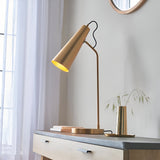 Endon Brockleigh antique brass table lamp with adjustable conical shade and black fabric flex, on a wooden console beside sheer curtains and a round mirror.