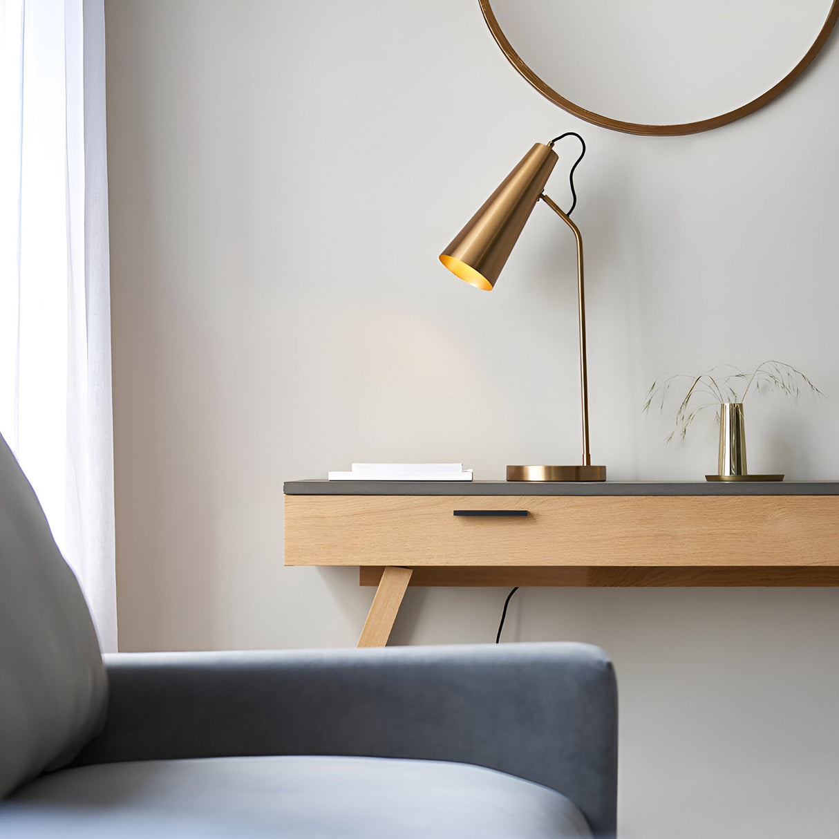 Endon Brockleigh antique brass table lamp with adjustable conical head on an oak console beneath a round mirror, beside a chair in a bedroom setting.