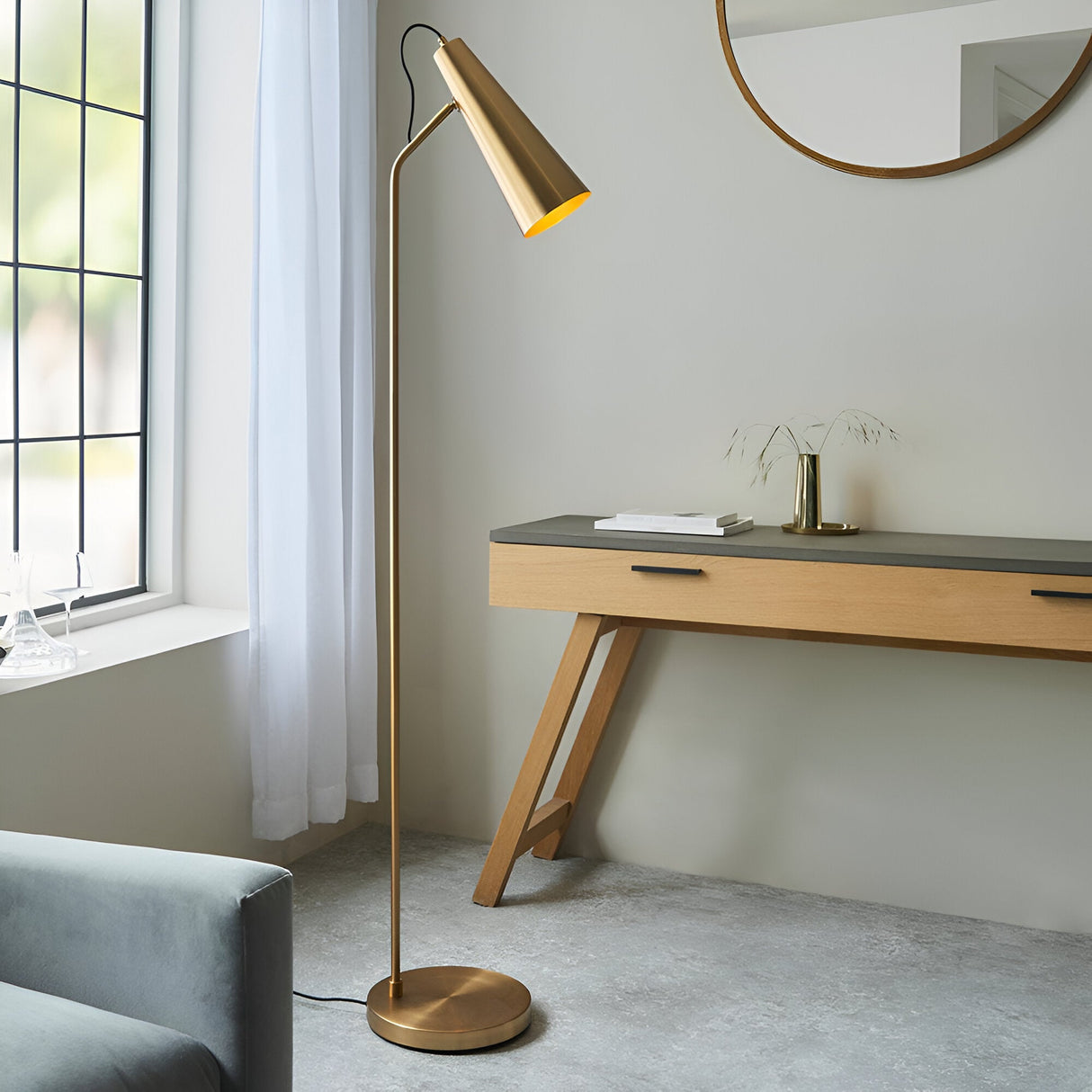 Brockleigh Task Floor Lamp in antique brass with an angled cone shade on a slender stem and round base, shown beside a console table in a living room.