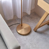 Brockleigh Task Floor Lamp in antique brass with a round base and slim pole, shown beside a sofa and wooden table leg on a grey floor.