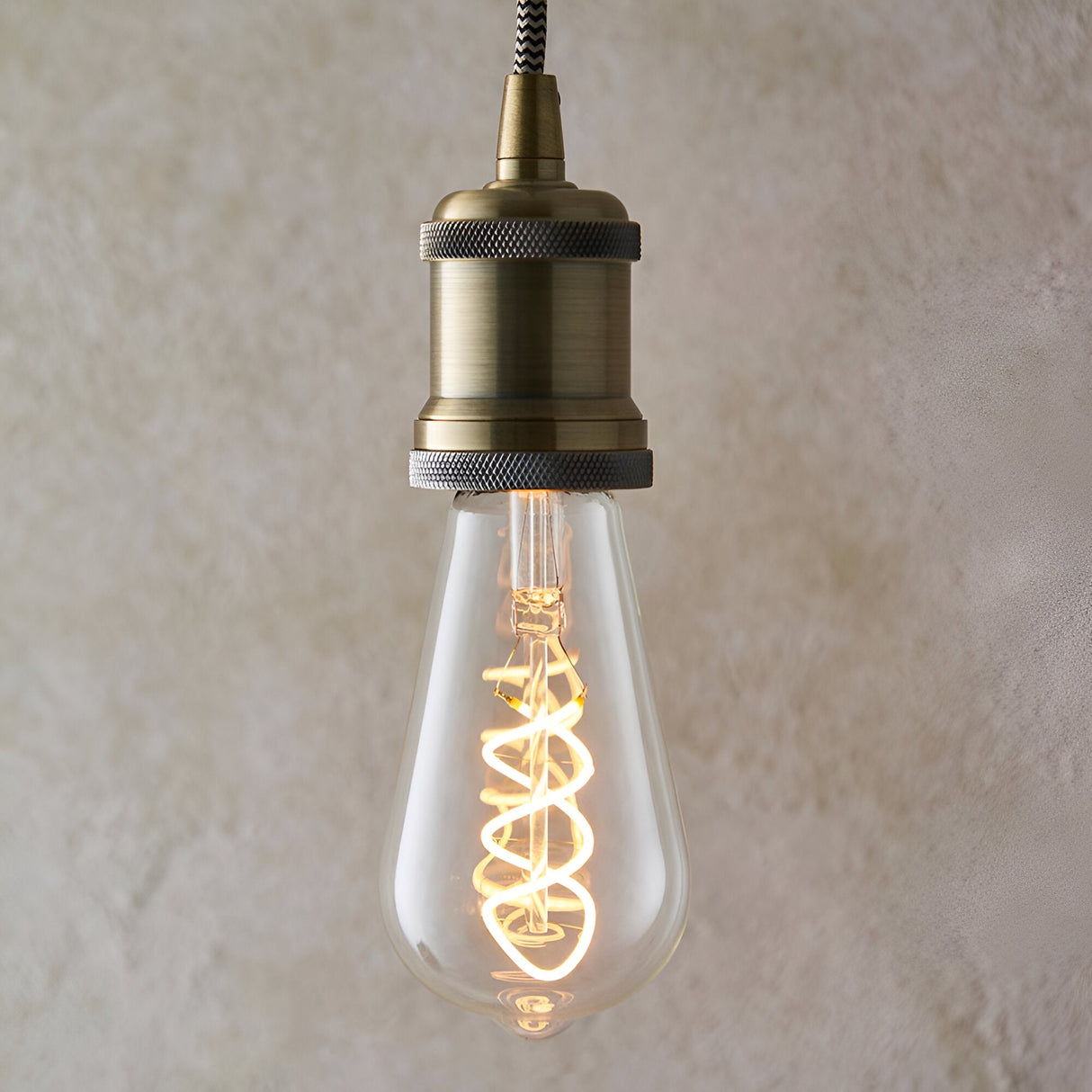 Endon 4W E27 LED filament twist bulb with clear teardrop glass and spiral filament, lit in warm white, suspended from an antique brass pendant holder.