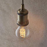 Antique brass E27 pendant holder with knurled detail, fitted with a clear GLS LED filament bulb glowing warm white, against a textured plaster wall.