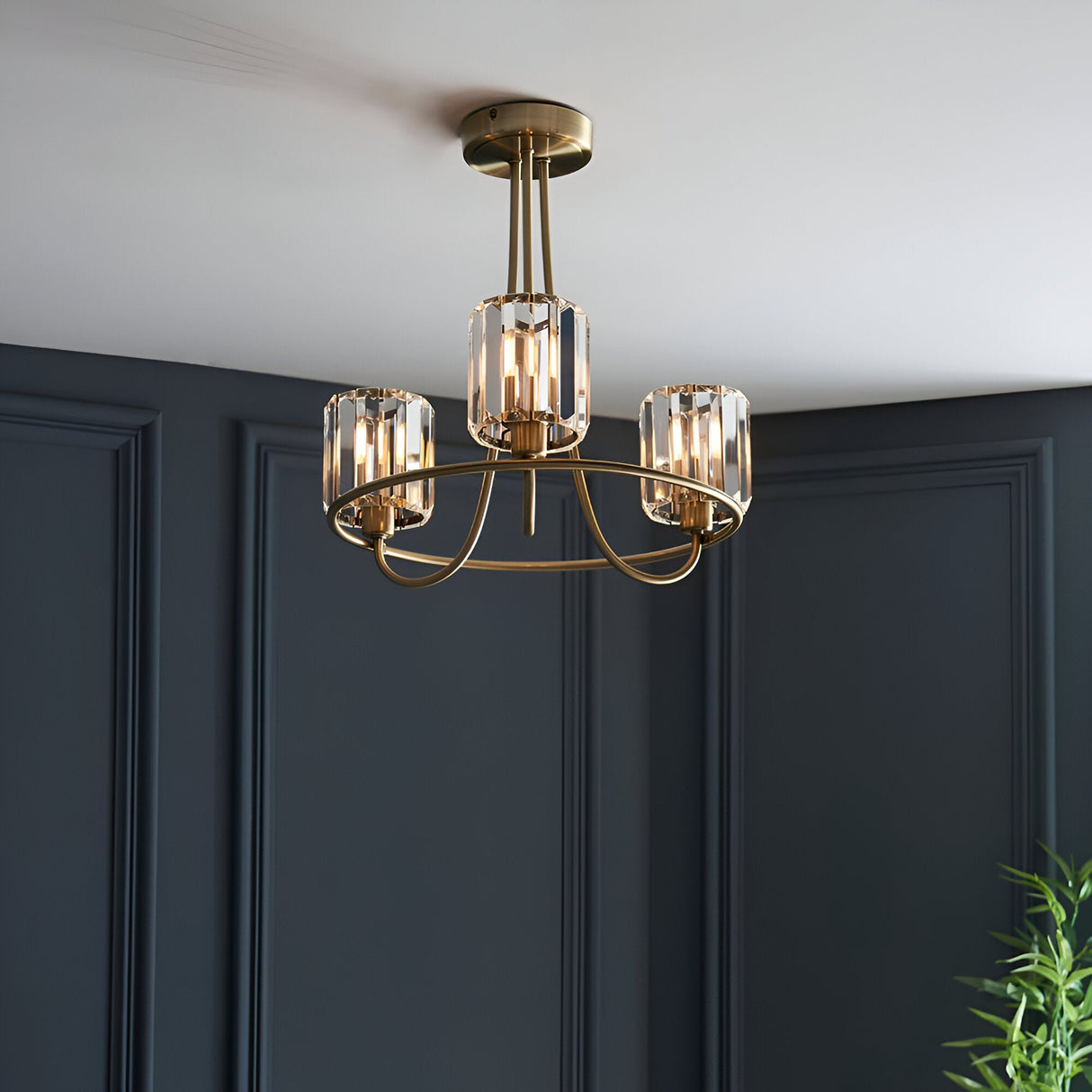 Endon Clementine 3 light semi-flush ceiling light in brass with faceted crystal glass shades, mounted below a white ceiling against a dark navy panelled wall.