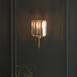 Endon Clementine wall light in brass with a cut faceted glass shade illuminated against a dark panelled wall.