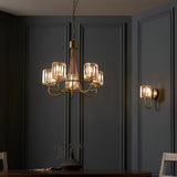 Endon Clementine brass wall light with faceted crystal glass shade, shown beside a matching chandelier on a dark grey panelled dining room wall.