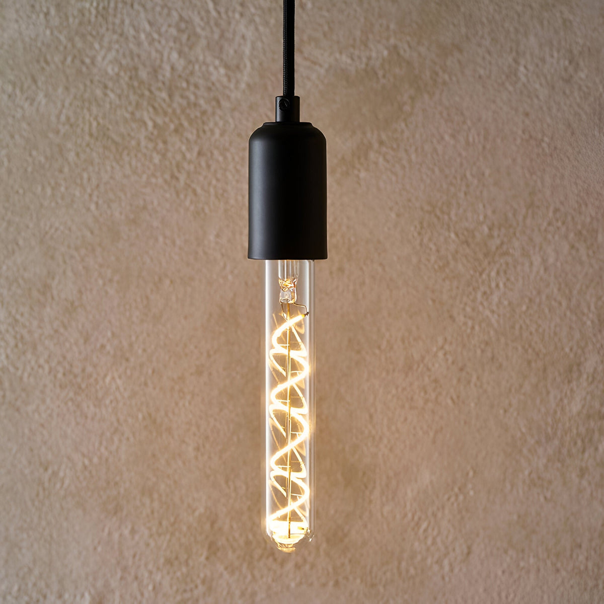 Endon 4W E27 LED filament bulb with clear tubular glass and warm white spiral filament, suspended from a matte black pendant holder against a beige wall.