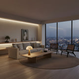 A modern living room features a beige sofa, two dark chairs, a round rug, and a wooden coffee table with Viren 2 Light Dome Crown Alabaster Table Lamps in Brushed Bronze & Soft White. Wall art and large windows highlight the city skyline at dusk.