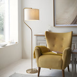 Bevmere Floor Lamp in aged brass with a curved stem, drum-shaped linen shade and round base, shown beside a mustard armchair in a living room.