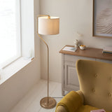 Bevmere Floor Lamp in aged brass with curved metal stem, drum-shaped linen fabric shade, and round base, shown beside a yellow armchair in a living room.