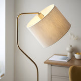 Bevmere Floor Lamp in aged brass with a curved metal stem and natural linen fabric drum shade, shown lit beside a desk in a living room setting.