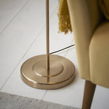 Close-up of the Bevmere Floor Lamp's round aged brass base and slender pole, shown beside a mustard armchair on a white wooden floor.