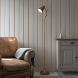 Endon Bevleigh brass floor lamp with flexible arm and single shade, lit beside a tan leather armchair and grey cabinet against panelled walls.
