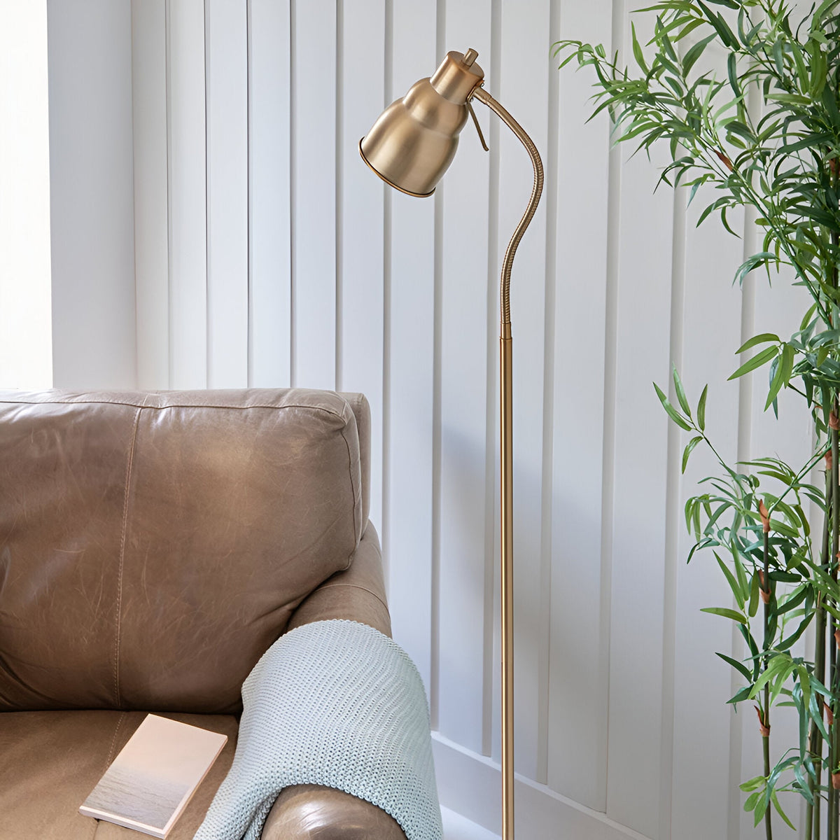Endon Bevleigh brass floor lamp with flexible gooseneck arm beside a tan leather sofa, styled with a knit throw and bamboo plant against panelled walls.