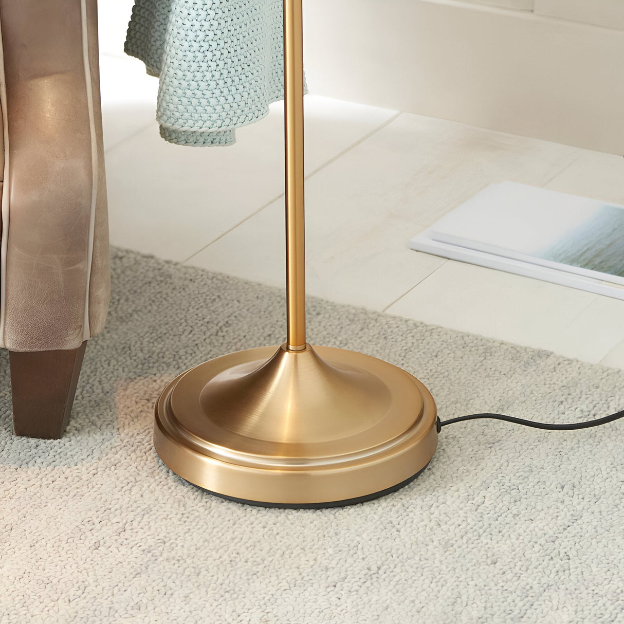 Close-up of the Endon Bevleigh floor lamp's round brushed brass base beside a leather armchair on a grey carpet in a living room setting.