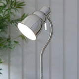 Endon Bevleigh floor lamp in nickel, close-up of the domed head and flexible gooseneck arm with rotary switch, set against a light panelled wall with foliage.