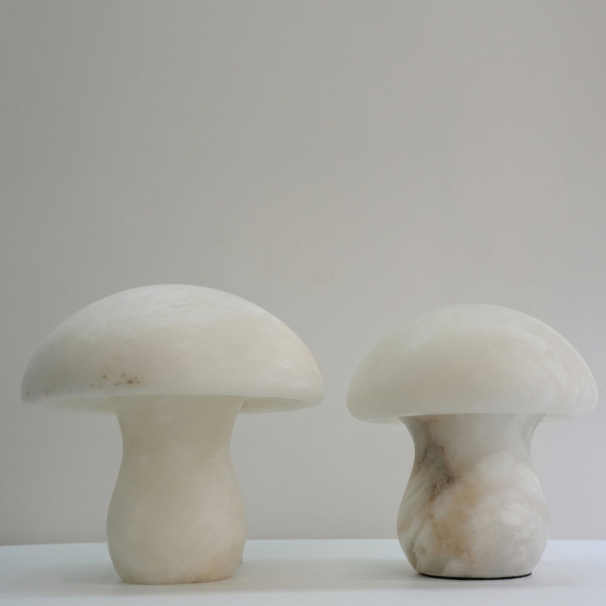 Two mushroom-shaped alabaster table lamps with domed caps and waisted bases in natural white and grey veined stone, shown side by side on a plain light background.