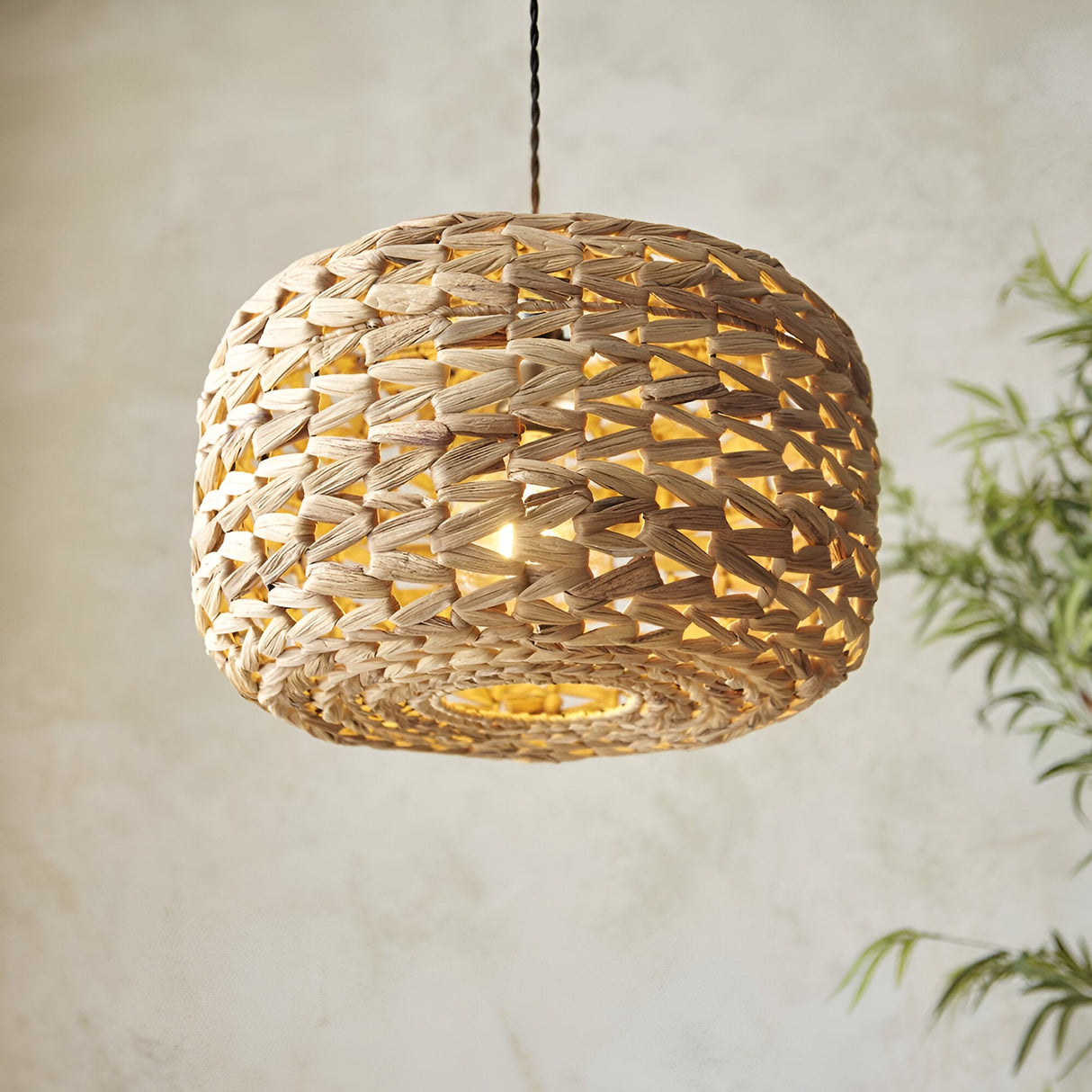 Endon Cranley pendant shade in natural handwoven rattan with a rounded drum shape, illuminated to show its open weave against a pale wall.
