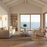 A bright, minimalist living room with light wood floors, a beige sofa, round coffee table, and ocean-view windows is softly illuminated by the Selvara LED Small Alabaster Double Wall Light in Brushed Brass & Soft White.