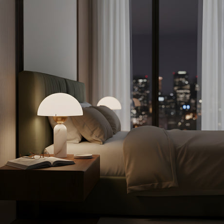 A modern bedroom at night features a neatly made bed and the Gildara 1 Light Dome Alabaster Table Lamp in Brushed Bronze & Soft White on a wooden nightstand with an open book, glasses, and coffee cup. City lights shine through large windows behind.