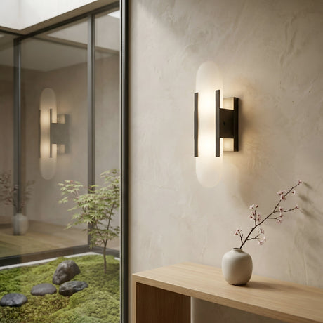 The Lirone 1 Light Small Vertical Capsule Alabaster Wall Light in soft white and black is mounted on a textured beige wall beside a minimalist wooden shelf with a vase of cherry blossoms. A glass wall offers views of a tranquil indoor garden with rocks and plants.