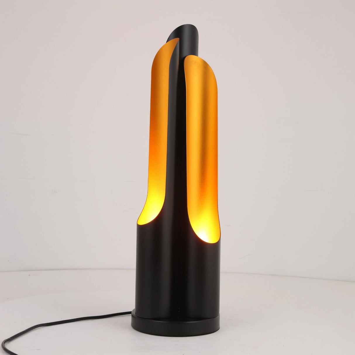 The Stealth Table Lamp - Brass & Black features curved panels in an abstract, sculptural design, partially enclosing a warm yellow light on a round base with a visible power cord.