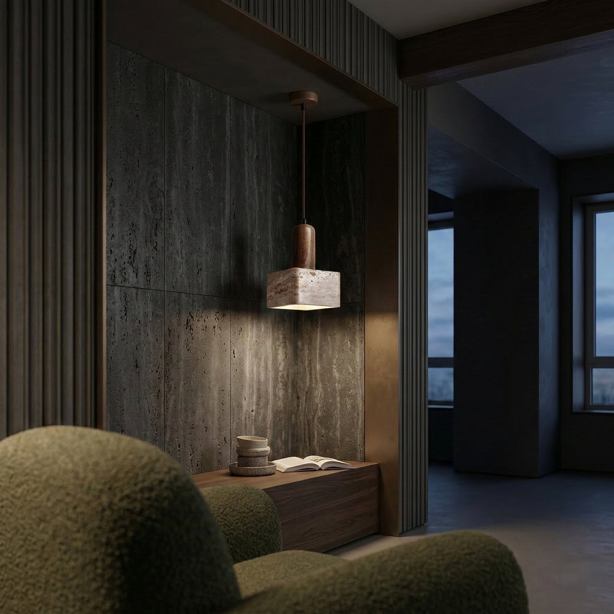 A cozy room features a textured green armchair, a wooden bench with a book and stacked bowls, illuminated by the Cavero 1 Light Square Block Travertine Single Pendant Light in Walnut Brown & Light Beige, against a dark textured wall and large windows at dusk.