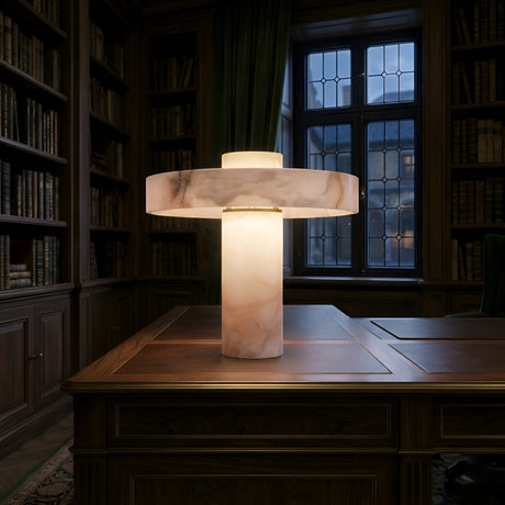The Zolara 1 Light Disc Alabaster Column Table Lamp in soft white glows atop a wooden desk in a dimly lit library, surrounded by bookshelves and a large window with a dark green curtain.