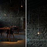 Berry S Aluminium LED pendant light with slim black tube and angled domed head, shown with another pendant against a stone wall in a dining room.