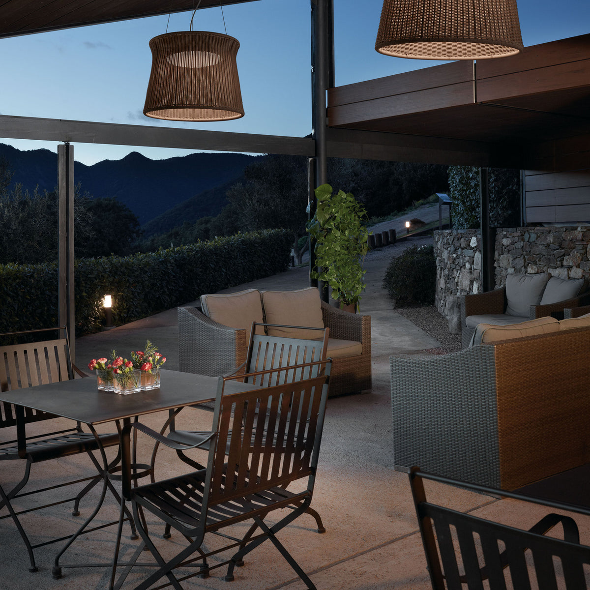 Syra 45 Aluminium LED pendant lights with woven brown drum shades hang above an outdoor dining and lounge patio overlooking mountains at dusk.