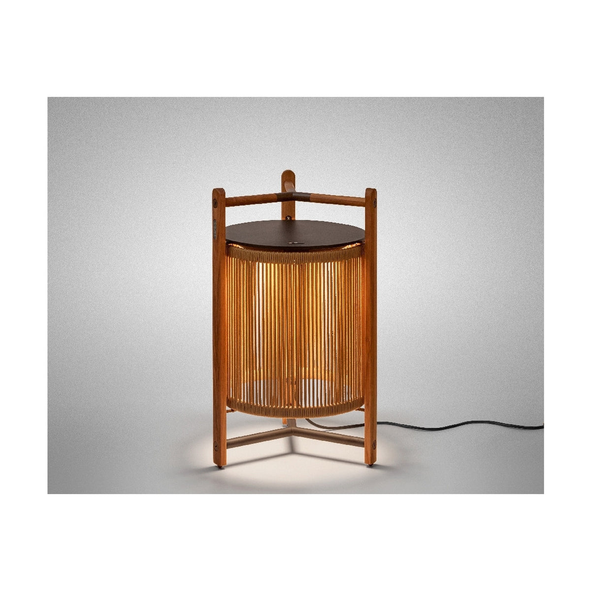 Kando P/76 outdoor floor lamp, lantern-style cylinder of sand nautical cord within a three-legged teak wood frame, dark round lid and top handle, lit, on a plain grey background.