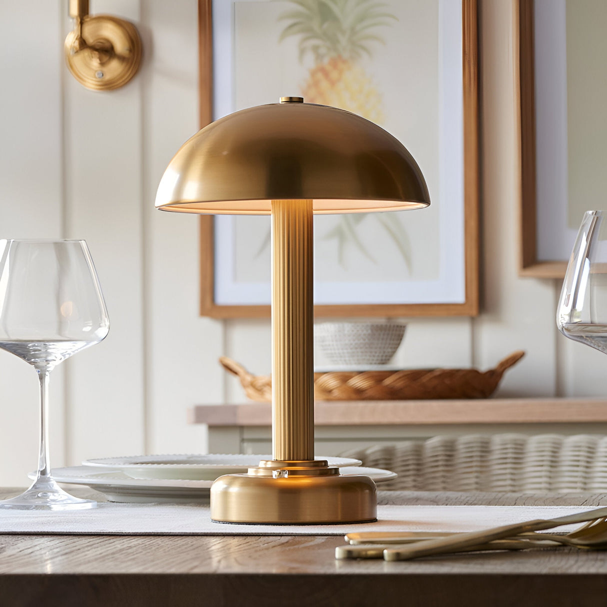 Endon Ashfield LED table lamp in brass with a domed shade and fluted stem, lit on a dining table beside wine glasses and framed pineapple art.
